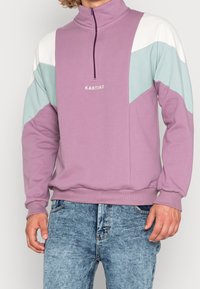 Fialová mikina s vysokým límcem, s barevně odstíněnými rukávy v světle modré a bílé barvě. Na přední straně je nápis "KAOTIKO".