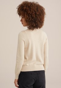 Svetlo bež pleten sweater z teksturiranim vzorcem, rebrastimi manšetami in robom. Nosi se z temnimi kavbojkami, kar prikazuje sproščen fit zadaj.