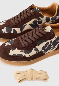 Adidași maro și crem cu un design imprimat cu vaci, accente din piele întoarsă, șireturi maronii și o talpă din cauciuc de tip gumă. Include un șiret suplimentar de crem.
