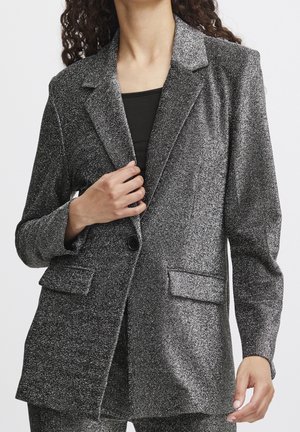 Kvinde iført en sort glitrende blazer med notchede revers og klaplommer, der holder blazeren lukket over en sort top.