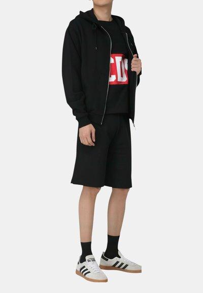 Osoba ubrana w czarną bluzę z zamkiem, czarne szorty, czarne skarpetki, białe trampki oraz czarną koszulkę z dużymi czerwono-białymi literami.