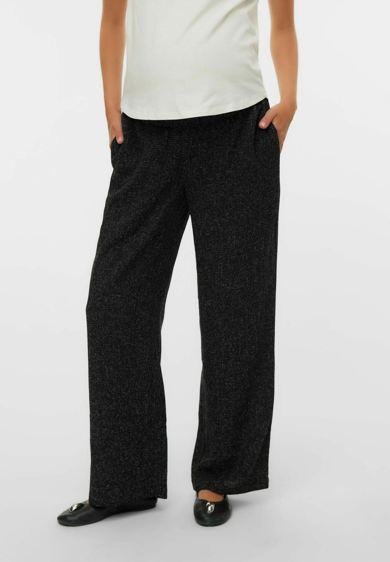 Hlače širokih hlačnic iz teksturirane črne tkanine z nežnimi pikčastimi vzorci. Imajo stranske žepe, kombinirane s svetlim zgornjim delom in črnimi čevlji.