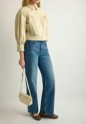 Kobieta w beżowej bluzce z guziczkami i bufkami na rękawach, niebieskich prostych jeansach, brązowych balerinach, trzymająca małą beżową torebkę za pasek.