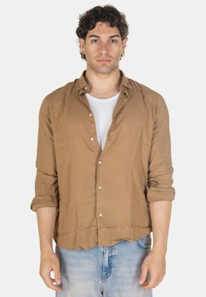 Uomo con capelli scuri e ricci indossa una camicia marrone con bottoni sopra una canotta bianca e jeans azzurri chiari, in piedi davanti a uno sfondo uniforme.