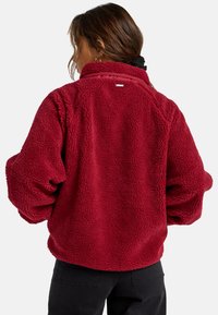 Giacca in fleece rossa con una superficie morbida e strutturata e colletto alto. Presenta spalle scese e una piccola etichetta sul retro. Sono indossati pantaloni neri.
