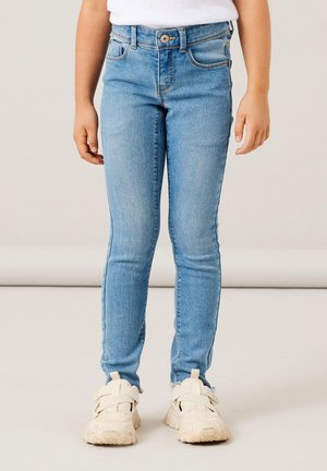 Jeans skinny in denim azzurro chiaro con chiusura frontale a bottone, passanti per cintura e design a cinque tasche, abbinati a sneakers beige.