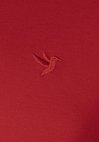 Rød tekstureret stof med et lille broderet kolibri-logo i en kontrastfarve, placeret mod den nederste højre hjørne.