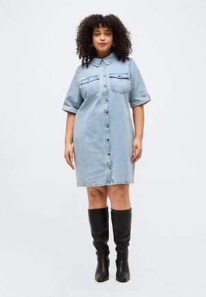 Ženska s kodrastimi lasmi v svetlo modri jeans obleki z gumbi in zavihanimi rokavi ter črnimi škornji do kolen, stoji pred belim ozadjem.