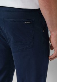 Donkerblauwe katoenen broek met een achterzak met label, gestructureerde pasvorm en een zachte textuur. Gedetailleerd met stiksels en een metalen knoop.
