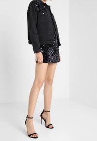 Musta farkkutakki, jossa on edessä napit ja kaksi taskua, kerroksittain mustan paljettimini-ompeluksen päällä. Yhdistetty mustien remmikenkien kanssa.