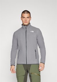 Grå fleece zip-up jakke med høj krave, logo på brystet og lynlåslommer på siden; har en blød tekstur og tætsiddende design.