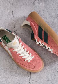Baskets en daim rose avec des bandes vert foncé, des lacets blancs, des accents crème et des semelles en gomme sur une surface en béton fissurée.