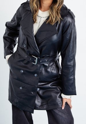 Kobieta ubrana w czarny skórzany płaszcz trencz z paskiem, nałożony na białą dzianinową bluzę i ciemne spodnie, stojąca z ręką w kieszeni.