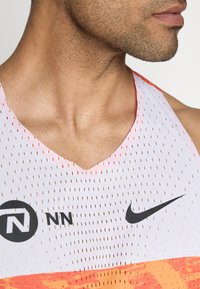 Valge sportlik tank-top perforeeritud kangast, mustade logode aktsentidega ning V-kaelusega disain. Heledast oranžist gradient allservas.