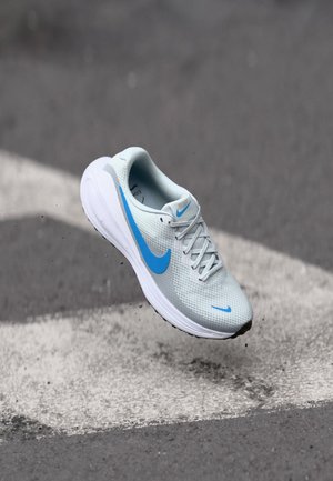Chaussure de course Nike gris clair et blanc avec un logo Swoosh bleu, en suspension au-dessus d'une route en asphalte avec des lignes blanches peintes et de petits débris en dessous.
