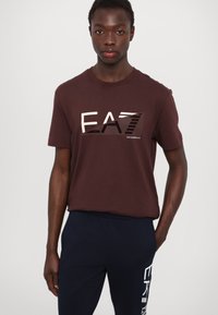 Barna rövid ujjú póló nagy "EA7" grafikával, különböző bézs árnyalatokban. Tengerészkék nadrággal párosítva, fehér felirattal az oldalon.