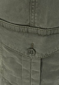 Detailní záběr šedé tkaniny s knoflíkem a šitím. Texturovaný povrch s jemným leskem. Viditelný detail kapsy.