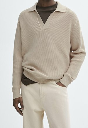 Mężczyzna w beżowym swetrze na drutach z dekoltem w serek, nałożonym na brązową koszulę i kremowe spodnie, stoi z jedną ręką w kieszeni na jednolitym tle.