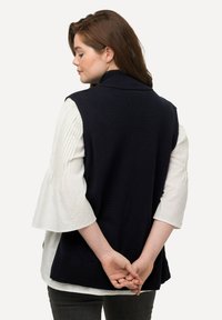 Gilet lavorato a maglia blu navy con colletto alto e orlo a coste, sovrapposto a una camicia bianca con maniche a campana plissettate.