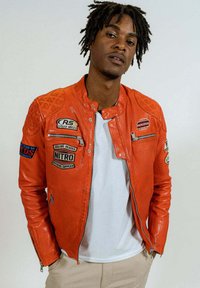 Veste en cuir orange avec des panneaux d'épaule texturés, plusieurs écussons et des poches zippées. Portée avec un t-shirt blanc en dessous.