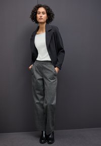 Veste noire, haut blanc et pantalon à carreaux gris. Le pantalon présente des détails de boutons sur le côté. Le mannequin se tient devant un fond gris.