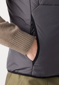 Grå pufferväst med svart kant, med en brun stickad ärm synlig. Vesten har en sidoficka för förvaring.