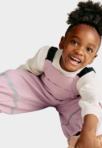 Salopettes roses imperméables avec des rayures grises, bretelles réglables et une poche avant, accompagnées d'un sweat-shirt blanc moucheté et de chaussures beiges.