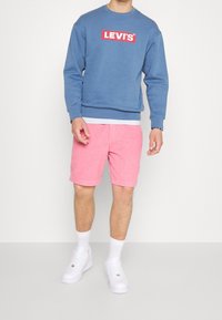 Persona che indossa una felpa blu Levi's, pantaloni shorts rosa, calzini bianchi e sneakers bianche, in piedi contro uno sfondo chiaro e uniforme.