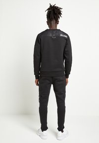 Černá mikina s lesklým zadním panelem, doplněná o sladěné kalhoty s bočními kapsami a bílé sportovní boty.