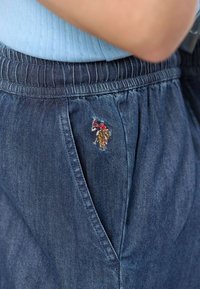 Pantaloni din denim cu nuanță albastru închis, având o talie elastică și un mic design brodat pe buzunar, evidențiind detalii și textură.