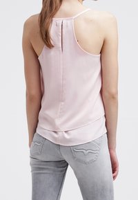 Haut sans manches rose clair avec un col rond et un détail de pli dans le dos, associé à un jean gris clair comportant des poches arrière décoratives.