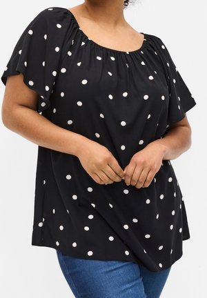 Woman wearing black short-sleeve blouse with white polka dots and gathered neckline, paired with blue jeans, hands clasped in front.