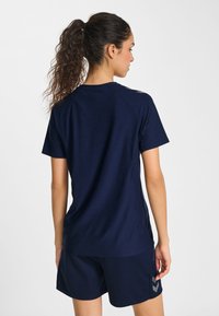 Femme avec queue de cheval bouclée portant un t-shirt de sport bleu marine et un short, vue de dos sur un fond blanc uni.
