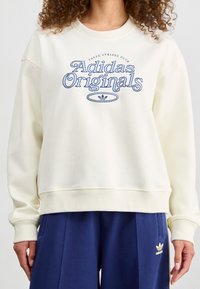 Krämfärgad sweatshirt med "Adidas Originals" grafik i blått, ribbad rundhalsning, lös passform och mjuk tygtextur. Matchas med marinblå byxor.