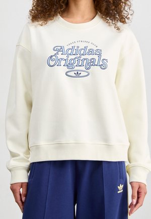 Sweatshirt de couleur crème avec un graphisme « Adidas Originals » en bleu, encolure ras du cou côtelée, coupe décontractée et texture de tissu douce. Associé avec un pantalon bleu marine.