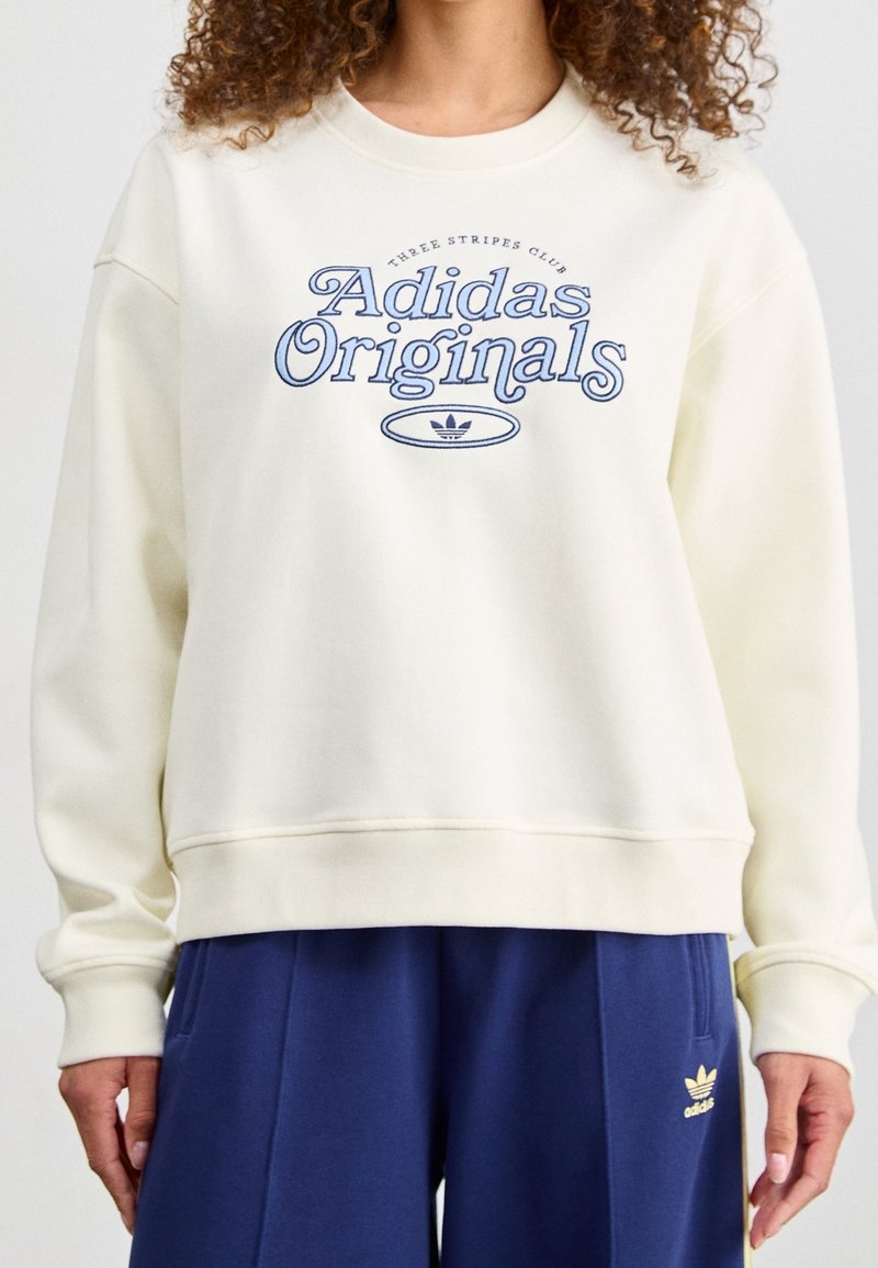 Krämfärgad sweatshirt med "Adidas Originals" grafik i blått, ribbad rundhalsning, lös passform och mjuk tygtextur. Matchas med marinblå byxor.