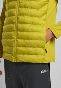 Leichte gelbe isolierte Jacke mit gestepptem Futter, durchgehendem Reißverschluss und Seitentaschen; kombiniert mit dunkelgrauen Hosen mit Logodetails.