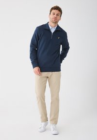 Tumesinise tõmblukuga pullover, millel on soonikkoes mansetid ja alläär, millel on väike kollane logo. Kombineeritud heledate beežide pükste ja valgete tossudega.