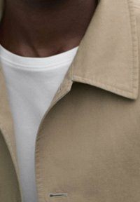 Cappotto beige con colletto, dotato di pattina per bottoni nascosta. T-shirt bianca sotto, visibile al colletto. Texture del tessuto lisce.