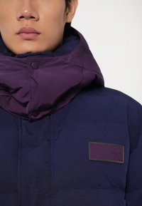 Modra puffer jakna s vijolično kapuco, z značilno prešito teksturo, zataknjenim zapenjanjem in majhnim pravokotnim logotipom v vijolični in zeleni barvi.