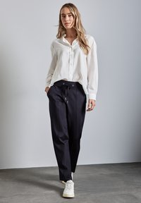 Camicia bianca a maniche lunghe in tessuto testurizzato, abbinata a pantaloni navy con coulisse. Il modello indossa sneaker bianche e si trova su uno sfondo grigio.