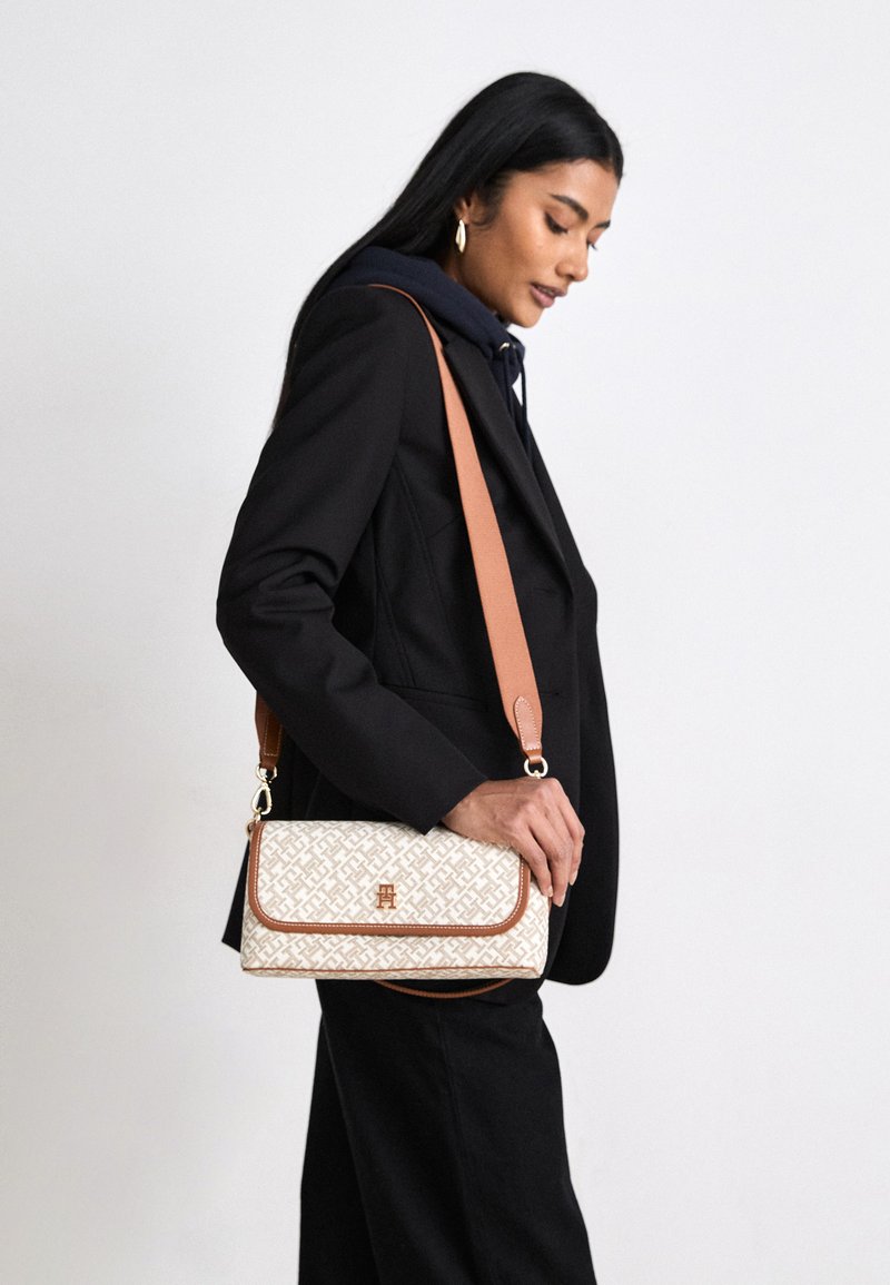 Bolsa crossbody com padrão branco e bege, com detalhes em couro castanho. Apresenta um fecho de aba e uma alça ajustável, segurada por um modelo com um blazer preto.