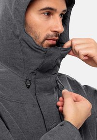 Giacca grigia con cappuccio, caratterizzata da una chiusura a toggle, una zip anteriore e un tessuto testurizzato. Mano che regola il toggle vicino all'area del mento.