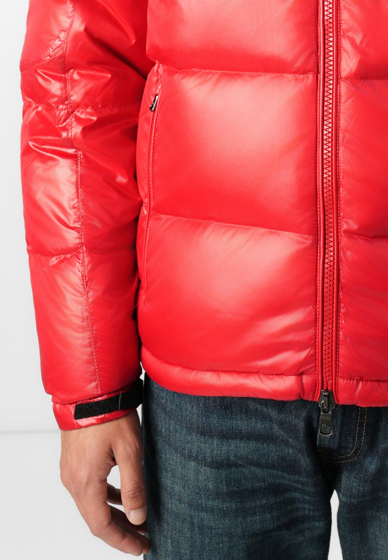 Rote Pufferjacke mit glänzendem Finish, gestepptem Design, Frontreißverschluss und verstellbaren Bündchen. Kombiniert mit Jeans.