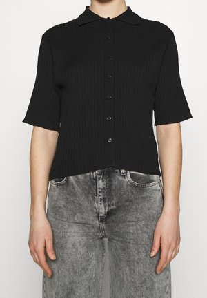 Vrouw draagt een zwart geribd, kort mouw knoopblouse gecombineerd met hoogwater, vervaagde grijze jeans, staand met ontspannen armen.