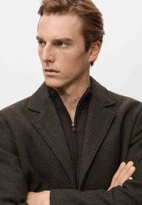 Abrigo de mezcla de lana color marrón oscuro con un cuello estructurado, que incluye una capa interior negra con cremallera, tela texturizada y una silueta estilizada.