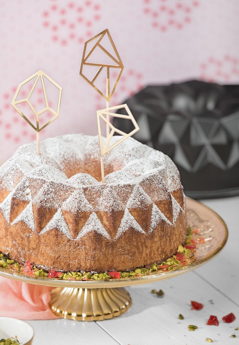 Bundt-Kuchen mit Puderzucker, geometrischen goldenen Kuchenfiguren, präsentiert auf einem goldenen Sockel mit Pistazien und Trockenfrüchten als Garnitur.