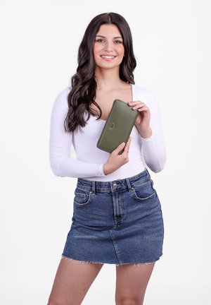 Green leather wallet with a zip closure, gold logo detail, and contrasting stitching. Model holding wallet, wearing a white top and denim skirt.