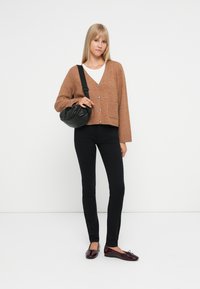 Cardigan marrone a maglia con tre bottoni e una tasca frontale, abbinato a pantaloni neri aderenti e ballerine bordeaux. Borsa a tracolla nera.