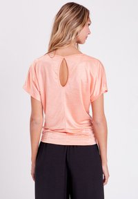 Haut à manches courtes couleur pêche avec un col rond et une ouverture en forme de clé à l'arrière ; fabriqué en matériau léger et lisse. Associé à un pantalon noir.